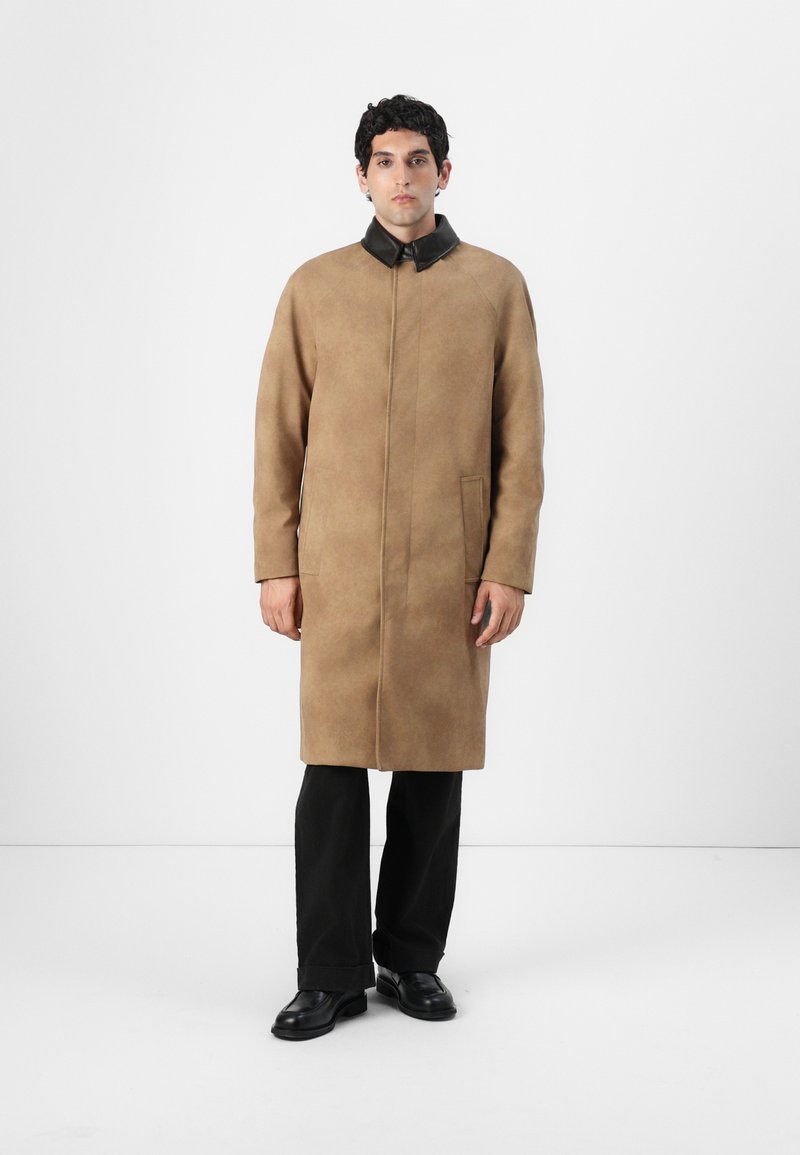 Manteau en daim tan avec un col en cuir noir, manches longues, et deux poches latérales, porté sur un pantalon et des chaussures noirs.