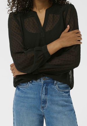 Femme aux cheveux bouclés portant un chemisier noir transparent à manches longues avec des pois et un jean bleu, les bras croisés sur la poitrine.