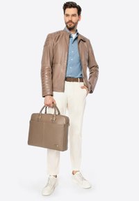 Giacca di pelle marrone con maniche testurizzate, camicia azzurro chiaro, pantaloni crema e sneakers bianche. Tiene una valigetta marrone coordinata.