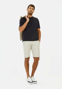 Marineblauwe t-shirt met korte mouwen, gecombineerd met lichtbeige shorts. Model houdt een lichtgrijze jas vast; draagt witte sneakers met donkere accenten.