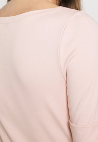 Světle růžové tričko s dlouhým rukávem, má ribbovanou texturu a široký výstřih. V oblasti ramen a na bocích jsou viditelné švy.