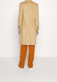 Cappotto in lana beige con taglio dritto, chiusura a doppio bottone e bottoni neri, abbinato a pantaloni color ruggine e sneaker casual.