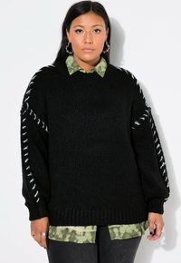 Schwarzer Strickpullover mit übergroßen Ärmeln, mit weißer Naht entlang der Schulterstücke. Trägt er über einem grünen Tie-Dye-Shirt.