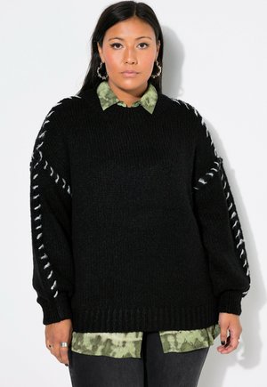 Schwarzer Strickpullover mit übergroßen Ärmeln, mit weißer Naht entlang der Schulterstücke. Trägt er über einem grünen Tie-Dye-Shirt.