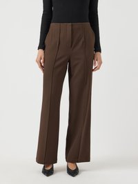 Mulher vestindo calças marrons de cintura alta e pernas largas com costuras frontais, combinadas com uma blusa preta de mangas longas e sapatos pretos de bico fino.