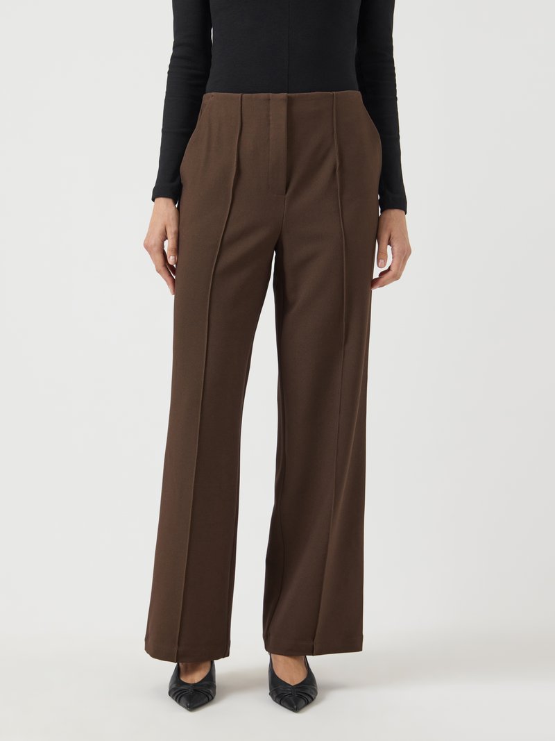 Mulher vestindo calças marrons de cintura alta e pernas largas com costuras frontais, combinadas com uma blusa preta de mangas longas e sapatos pretos de bico fino.