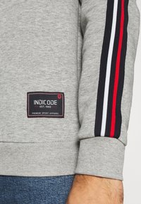 Sweatshirt gris avec des rayures rouges, noires et blanches sur les manches. Comprend une étiquette noire avec "INDICODE EST. 1993" et "VÊTEMENTS DE SPORT PREMIUM."