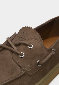 Καφέ σουέτ μοκασίνι με δερμάτινα κορδόνια που περνούν μέσα από ασημένια ροδέλες και υφασμένη σόλα, με λεπτομερείς ραφές και ανάγλυφο λογότυπο.