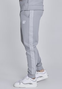 Svetlo sivé tepláky vyrobené z mäkkej tkaniny, so slim fit strihom, bočnými vreckami a vertikálnym vzorom pozdĺž švov s bielym brandingom.