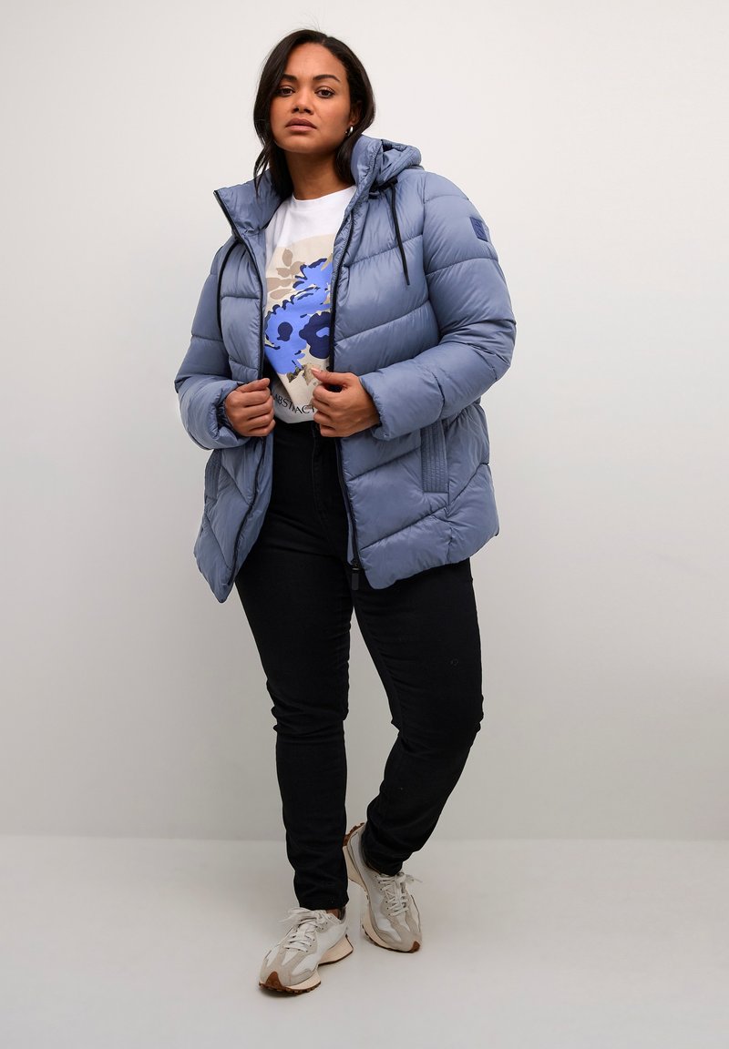 Femme portant une veste rembourrée bleu clair, un t-shirt graphique blanc, un pantalon noir et des baskets blanches, se tenant devant un fond uni clair.