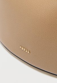 Beež tekstureeritud nahast kott, millel on kuldne logoaksent ja sile must servaääris. Pinnal on peen tera muster.