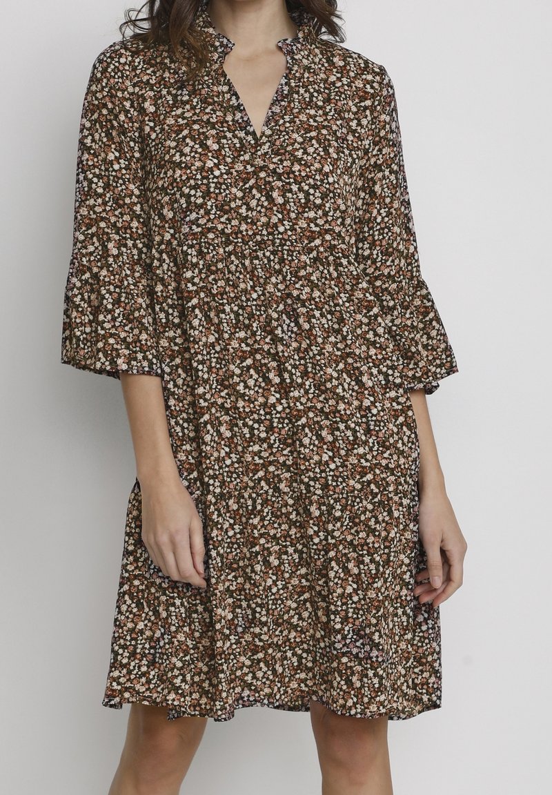 Femme portant une robe fleurie ample mi-longue avec des manches trois-quarts et un petit col à volants sur un fond clair uni.