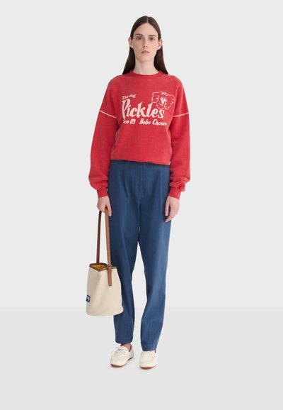 Femme portant un sweat-shirt rouge avec texte et graphisme, pantalon taille haute bleu, baskets blanches, tenant un sac seau crème avec bandoulière marron.