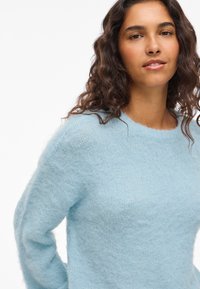 Svetlomodro teksturirano pulover z okrolim izrezom, dolgimi rokavi in mehkim, puhastim videzom. Ima udoben kroj in subtilen vzorec pletenine.