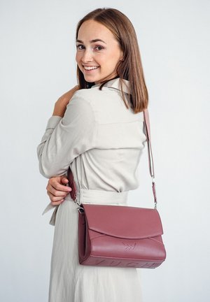 Burgunderfarbene Lederhandtasche mit Klappenverschluss, geprägtem Logo, abnehmbarem Tragegurt und strukturierter Form. Das Modell hält sie über der Schulter.