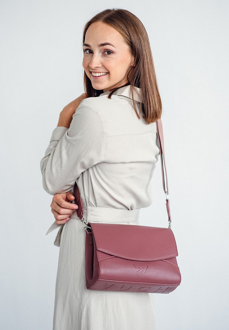 Burgunderfarbene Lederhandtasche mit Klappenverschluss, geprägtem Logo, abnehmbarem Tragegurt und strukturierter Form. Das Modell hält sie über der Schulter.