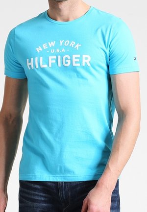 Mężczyzna w jasnoniebieskiej koszulce z napisem "NEW YORK U.S.A HILFIGER" w białym kolorze, w połączeniu z ciemnoniebieskimi dżinsami.