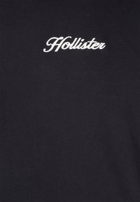 Svart bomullströja med vit broderad "Hollister"-text, med en mjuk textur och design med rund hals.