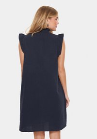 Rochie fără mâneci de culoare bleu marin, cu accente volanate la umeri, croi lejer, țesătură texturată și tiv drept. Dispune de o cusătură centrală pe spate.