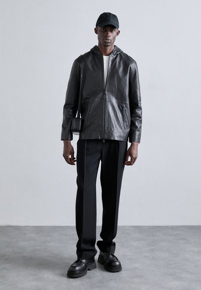 Jaqueta de couro preta com fecho, capuz, combinada com uma camisa branca, calças pretas ajustadas e sapatos pretos robustos. Inclui uma pequena bolsa transversal.