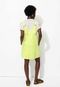 Robe salopette en denim jaune avec des bretelles réglables, des poches latérales et une coupe décontractée, portée par-dessus un T-shirt beige clair.