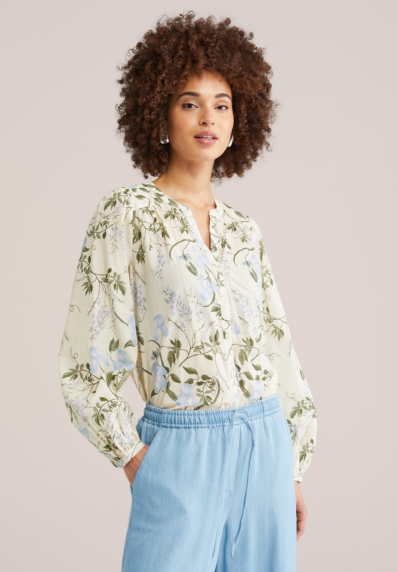 Kvinde med krøllet hår iført en cremefarvet blomsterbluse med blå accenter og lyseblå bukser med snøre mod en ensfarvet baggrund.