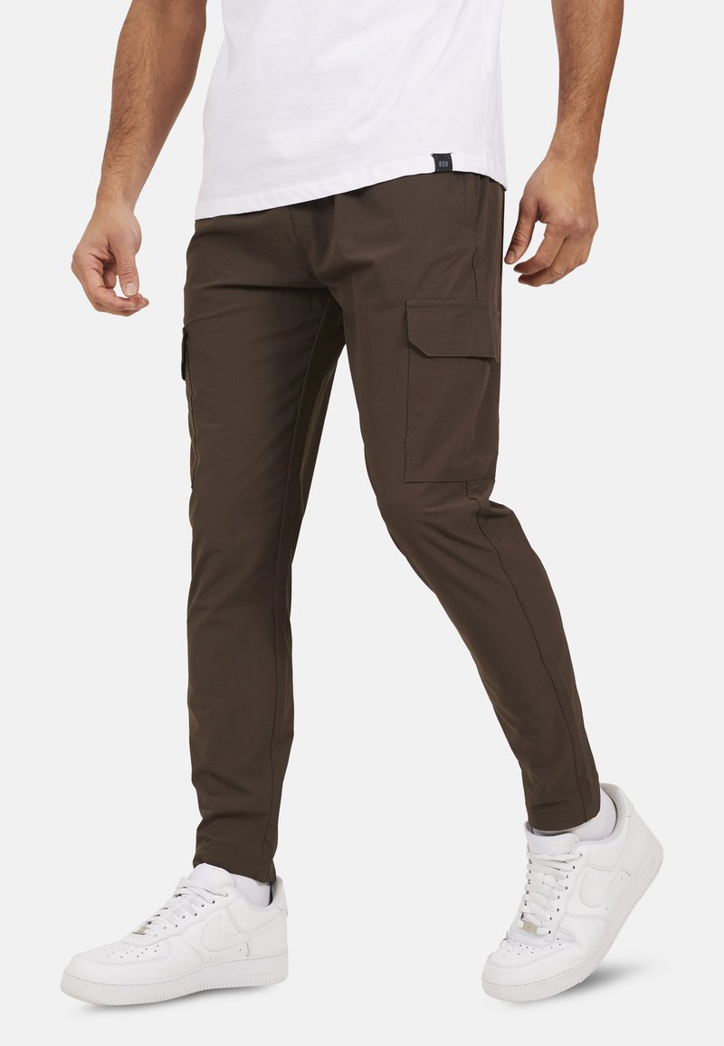 Brązowe spodnie cargo o slim fit, z bocznymi kieszeniami i gładką fakturą, w połączeniu z białymi trampkami i prostą białą koszulką.