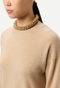 Collier ras du cou doré à perles avec plusieurs brins texturés, porté sur un pull beige clair, présentant une surface lisse et des accents de perles rondes.