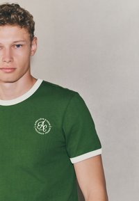 T-shirt verde in cotone con collo e polsini bianchi a coste. Presenta un logo circolare sul lato sinistro del petto. Design semplice.