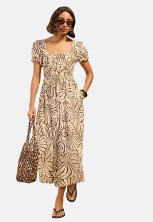 Kvinde iført beige og brun blomsterprint midi-kjole med puffede ærmer, solbriller, sandaler, armbånd og bærer en flettet taske.