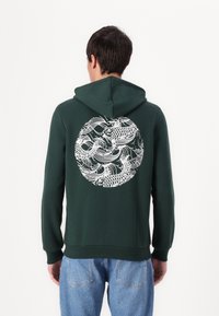 Dunkelgrüner Kapuzenpullover mit einem weißen, kreisförmigen Design mit Fisch- und Wellenmustern auf der Rückseite. Mit Kängurutasche und Kordelzugkapuze.