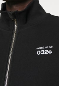 Veste noire zippée avec un col montant et le texte blanc "SOCIÉTÉ DE 032c" sur la poitrine gauche.