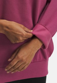 Fioletowy sweter z okrągłym dekoltem, ściągaczami na rękawach i gładką fakturą. Ręka dostosowuje mankiet, podkreślając elastyczność materiału.