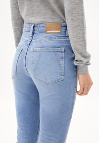 Světle modré džíny z denimu s přiléhavým střihem, vybavené dvěma zadními kapsami a hnědou značkovou nášivkou na pasu. Měkká textura.