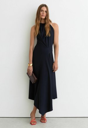 Nő ujjatlan fekete midi ruhában, kezében sötét kis táskával, arany karkötőt és piros pántos magassarkú cipőt visel, fehér fal előtt áll.