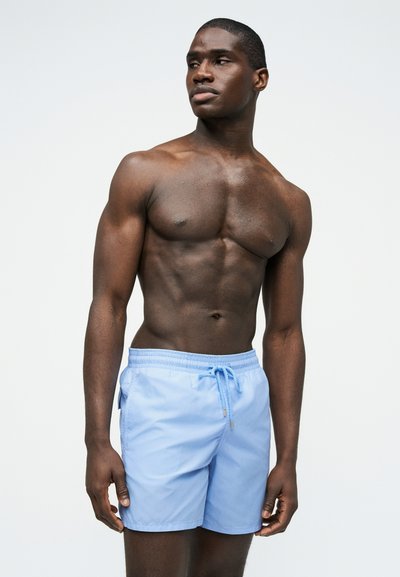 Μυώδης άνδρας με σκούρο δέρμα που φοράει ανοιχτό μπλε μαγιό, στέκεται και κοιτάζει προς τα πλάγια σε απλό ανοιχτόφωτο φόντο.