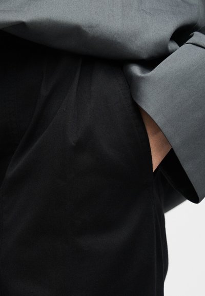 Άτομο που φοράει μαύρο παντελόνι με το χέρι μέσα στην τσέπη, ντυμένο με γκρι μακρυμάνικη μπλούζα με εμφανές το μανίκι.