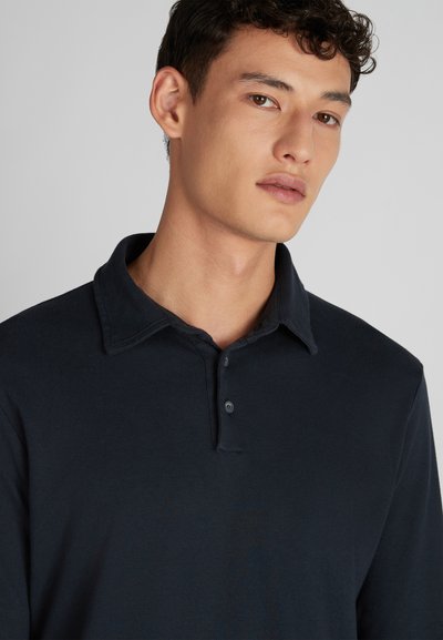 Un jeune homme aux cheveux bouclés courts portant une chemise à col boutonnée bleu marine, regardant légèrement sur le côté contre un fond uni.