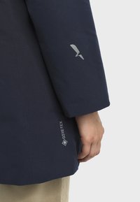 Desna ruka i ruka osobe koja nosi tamnoplavu Gore-Tex jaknu s logotipom na izlogu i donjem dijelu kaputa, vidljive bež hlače.