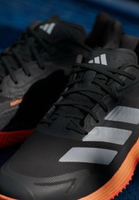 Μαύρα αθλητικά παπούτσια με υφασμάτινο επάνω μέρος με υφή, που διαθέτουν τρεις λευκές ρίγες, μαύρα κορδόνια και πορτοκαλί ελαστική σόλα. Κοντινή θέα.