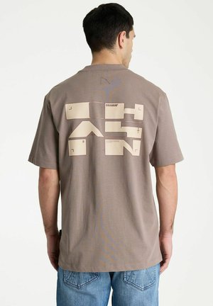 Man draagt een taupekleurig T-shirt met beige geometrisch blokontwerp en tekst op de achterkant, gecombineerd met blauwe spijkerbroeken, staand tegen een witte achtergrond.