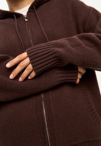 Sudadera con cremallera de punto marrón que presenta puños y dobladillo de canalé, con un acabado texturizado y un detalle de bolsillo tipo canguro.
