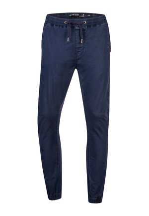 Homme portant un pantalon de jogging bleu marine avec taille à cordon et poignets élastiques, associé à des baskets blanches Reebok.