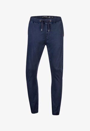 Homme portant un pantalon de jogging bleu marine avec taille à cordon et poignets élastiques, associé à des baskets blanches Reebok.
