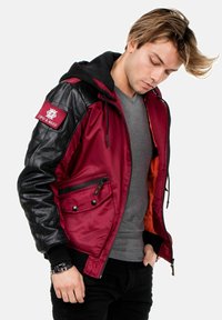 Rote und schwarze Bomberjacke mit Kapuze, mit gestepptem Lederärmel, Reißverschlusstasche, zwei Frontknöpfen und orangefarbener Innenausstattung.