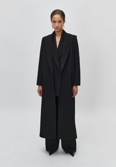 Donna che indossa un lungo cappotto nero su misura sopra un completo nero con pantaloni a gamba larga e scarpe nere con punta, in piedi contro uno sfondo semplice.