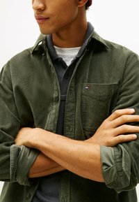 Olijfgroen corduroy shirt met knoopsluiting aan de voorkant, borstzak en logo-accent. Voorzien van opgerolde mouwen en een gestructureerde afwerking over een grijze polo.