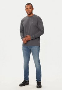 Camiseta de manga larga gris oscuro con cuello redondo y pequeño logo en el pecho, combinada con jeans de mezclilla azul claro y zapatillas negras.