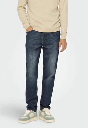 Donkerblauwe denim jeans met een recht model, gedragen met een lichtbeige gebreide trui met patroon en grijze sneakers met witte accenten.