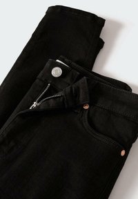 Mustat farkut, taitettuina, joissa on hopeanvärinen nappi, osittain auki oleva vetoketju ja näkyvä etutaskujen ompelu valkoista taustaa vasten.